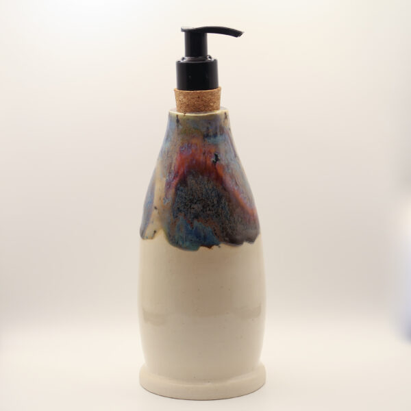 Tall ceramic soap bottle in cream glaze with blue, red and copper drips around the top, black pump in a cork stopper