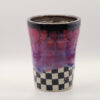 Handmade ceramic cup with a dark brown rim, drippy pink and purple glaze, and a black and white checkered base