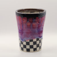 Handmade ceramic cup with a dark brown rim, drippy pink and purple glaze, and a black and white checkered base