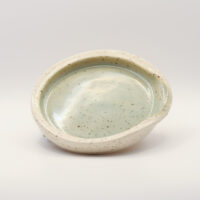 Shallow speckled spoon rest with a creamy white rim and pale sage green glazed centre, with a notch in the rim for a handle