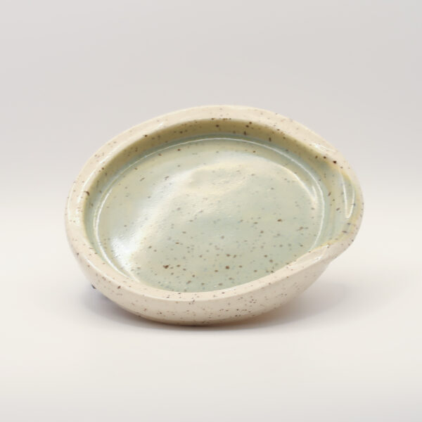 Shallow speckled spoon rest with a creamy white rim and pale sage green glazed centre, with a notch in the rim for a handle