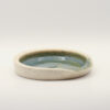 Shallow round spoon rest in speckled cream clay with a glossy blue-green glazed interior and a notch in the rim