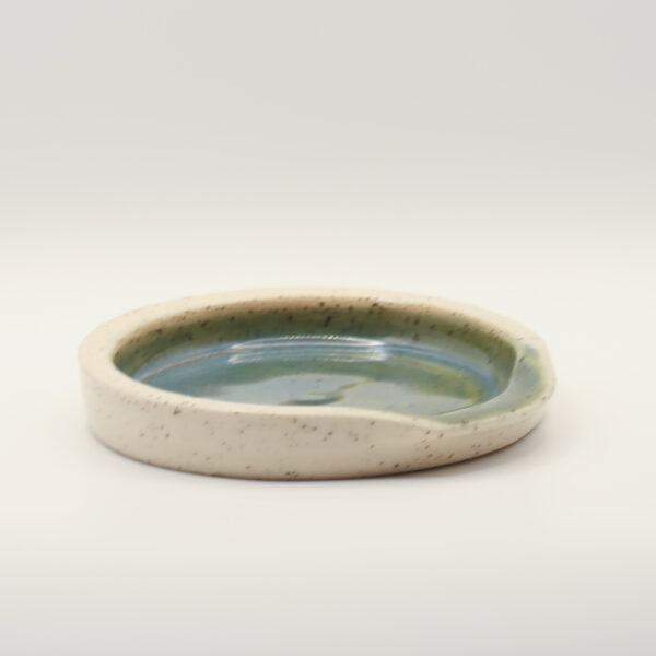 Shallow round spoon rest in speckled cream clay with a glossy blue-green glazed interior and a notch in the rim