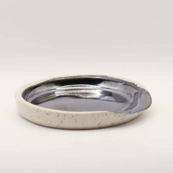Shallow round spoon rest with a glossy metallic black-silver glazed interior and a speckled cream exterior and rim