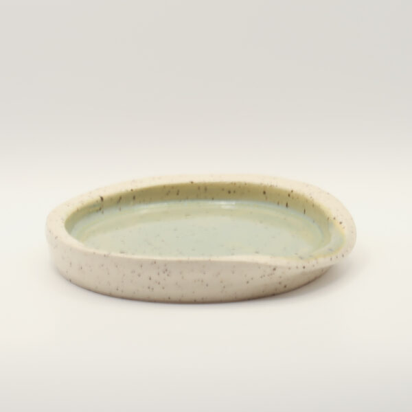 Shallow speckled cream stoneware spoon rest with a pale green glazed interior and a notch in the rim, on a white background