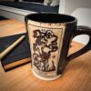 img_7887 Dark brown glazed mug with cream panel showing a Death tarot card skeleton, beside a black notebook and pen on a table