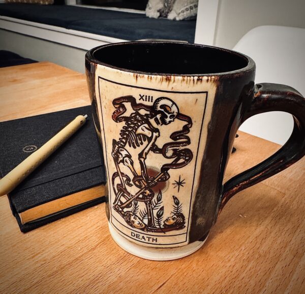 img_7887 Dark brown glazed mug with cream panel showing a Death tarot card skeleton, beside a black notebook and pen on a table
