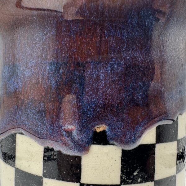 img_7929-2 Close-up of a ceramic cup with a drippy purple, red and blue glaze flowing over a black and cream checkerboard band